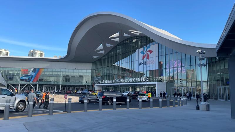 Exterior view of the Las Vegas Convention Center's West Hall under a clear blue sky. A large 'AHR Expo 2024' banner is displayed on the glass facade, with cars and people moving near the entrance